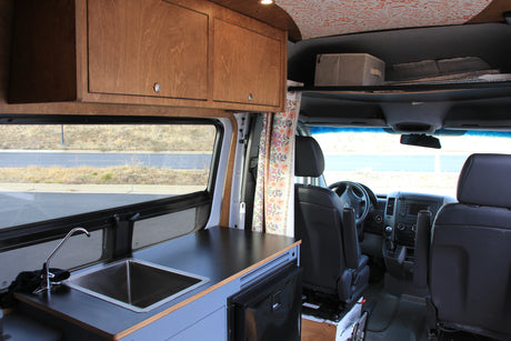 This RV rental features a camper van interior with a kitchenette, wooden overhead cabinets, and a view into the driver's cabin.