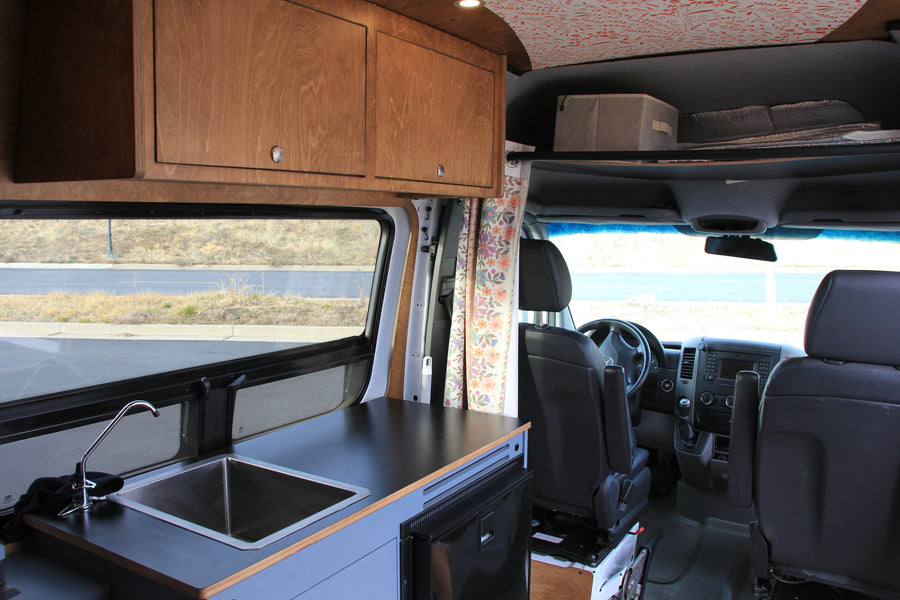This RV rental features a camper van interior with a kitchenette, wooden overhead cabinets, and a view into the driver's cabin.