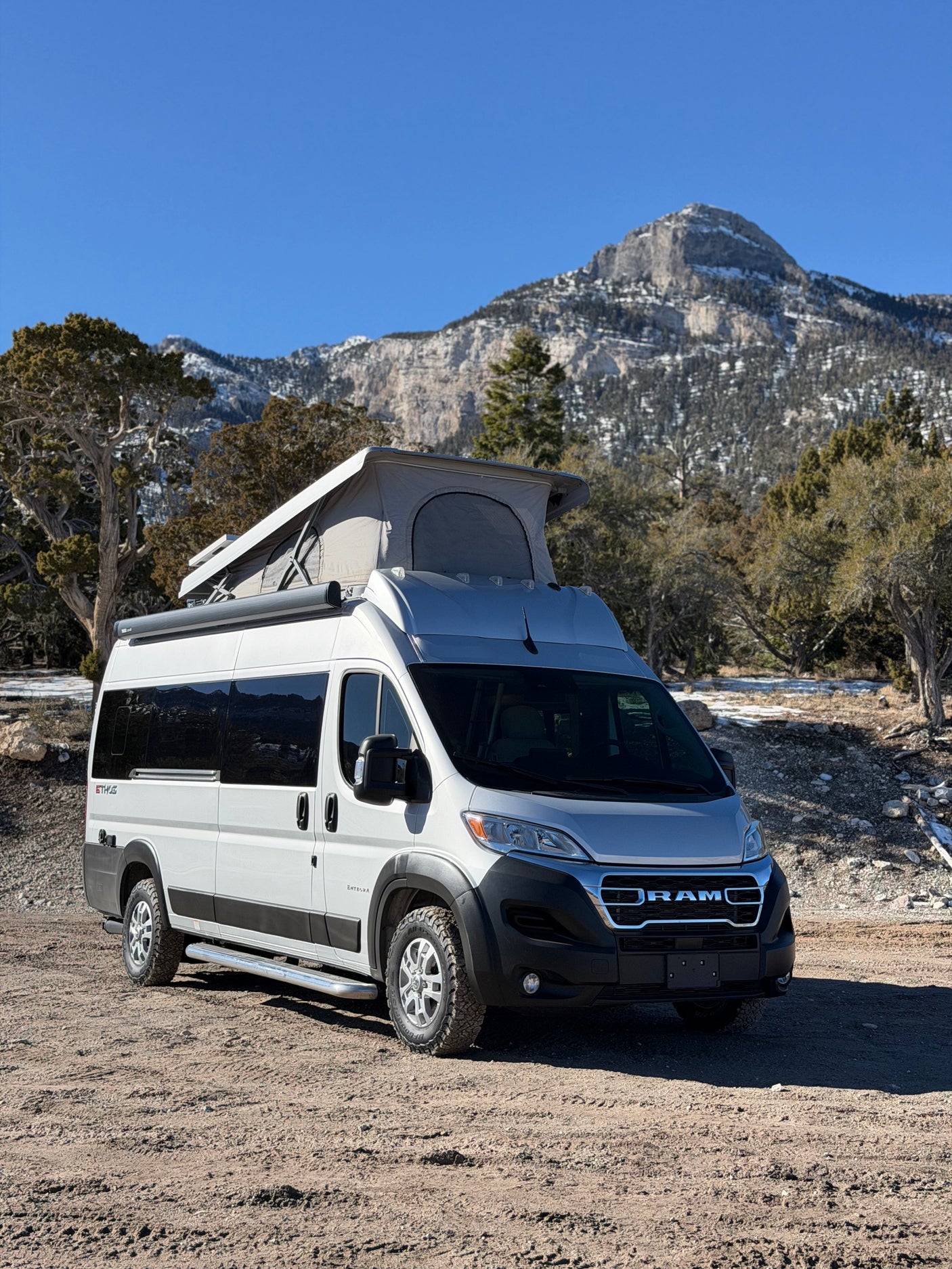 Experience mountain adventures with this rv rental camper van, featuring a pop-top, parked on dirt with snow-capped peaks and trees.