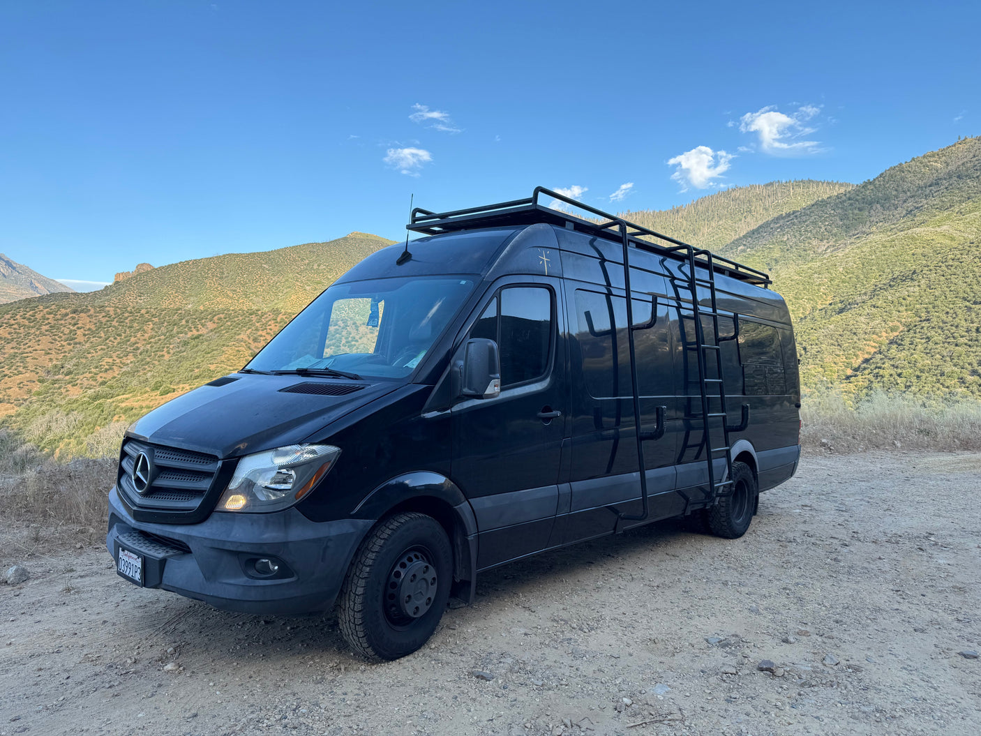 Black Mercedes Sprinter camper van, equipped for adventure with roof rack & ladder, parked on a dirt road amidst hills. Ideal RV rental.