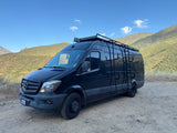 Black Mercedes Sprinter camper van, equipped for adventure with roof rack & ladder, parked on a dirt road amidst hills. Ideal RV rental.