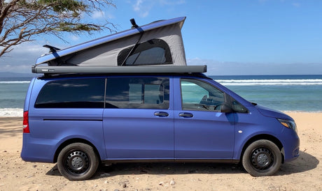 Blue Mercedes-Benz camper van with pop-top roof on a sandy beach. Perfect for your next RV rental & ocean adventures.