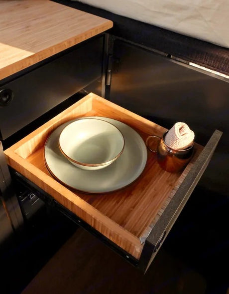 Inside an RV rental, a wooden drawer holds light green dishes with gold trim & a copper mug with a small towel.
