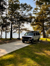 White RV rental van at a sunny lakefront campsite with trees. Ideal for outdoor travel and nature.