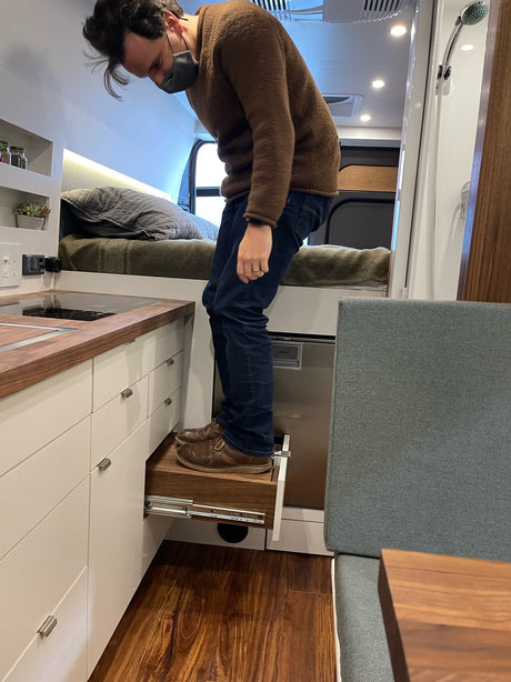 Man on pull-out step inside an RV rental. Smart compact design with kitchen, bed, and shower for mobile living.
