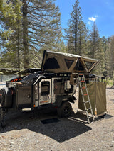 This rugged overland RV rental trailer features a rooftop tent, ladder, and privacy shower in a forest setting, ideal for camping adventures.