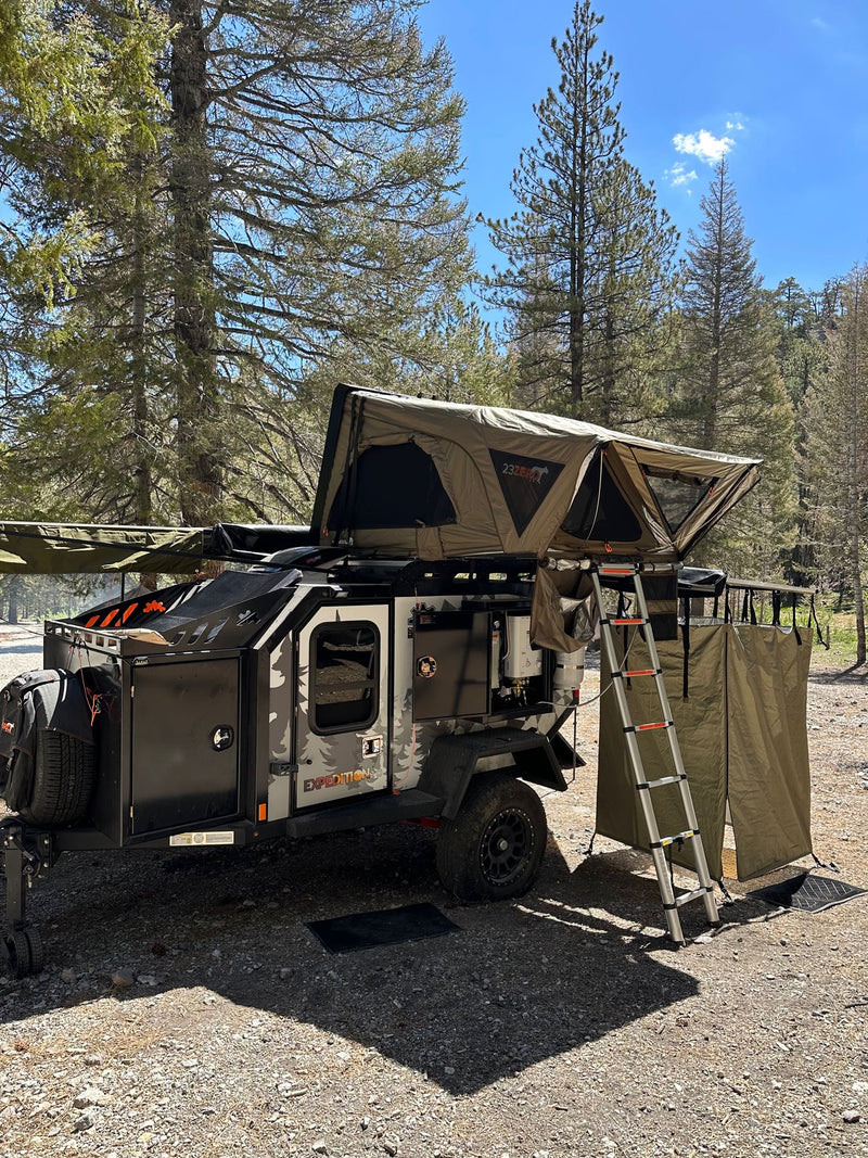 This rugged overland RV rental trailer features a rooftop tent, ladder, and privacy shower in a forest setting, ideal for camping adventures. — photo 15