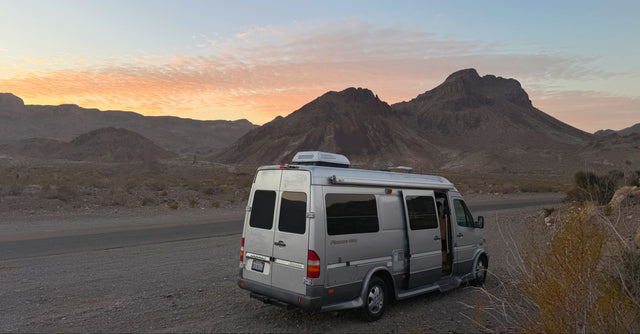 Silver RV rental camper van, a Pleasure-Way, parked by a desert road at sunrise among mountains, ready for adventure.