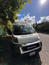 Bright Winnebago Ram ProMaster rv rental van parked under a tree on a sunny, cloudy day. Reflections in chrome grill, CA plate.