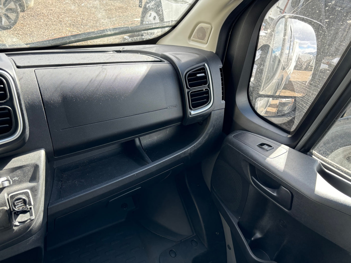 Inside an RV rental: passenger dashboard with vents, storage, and door controls. A dusty windshield shows the exterior. — photo 13