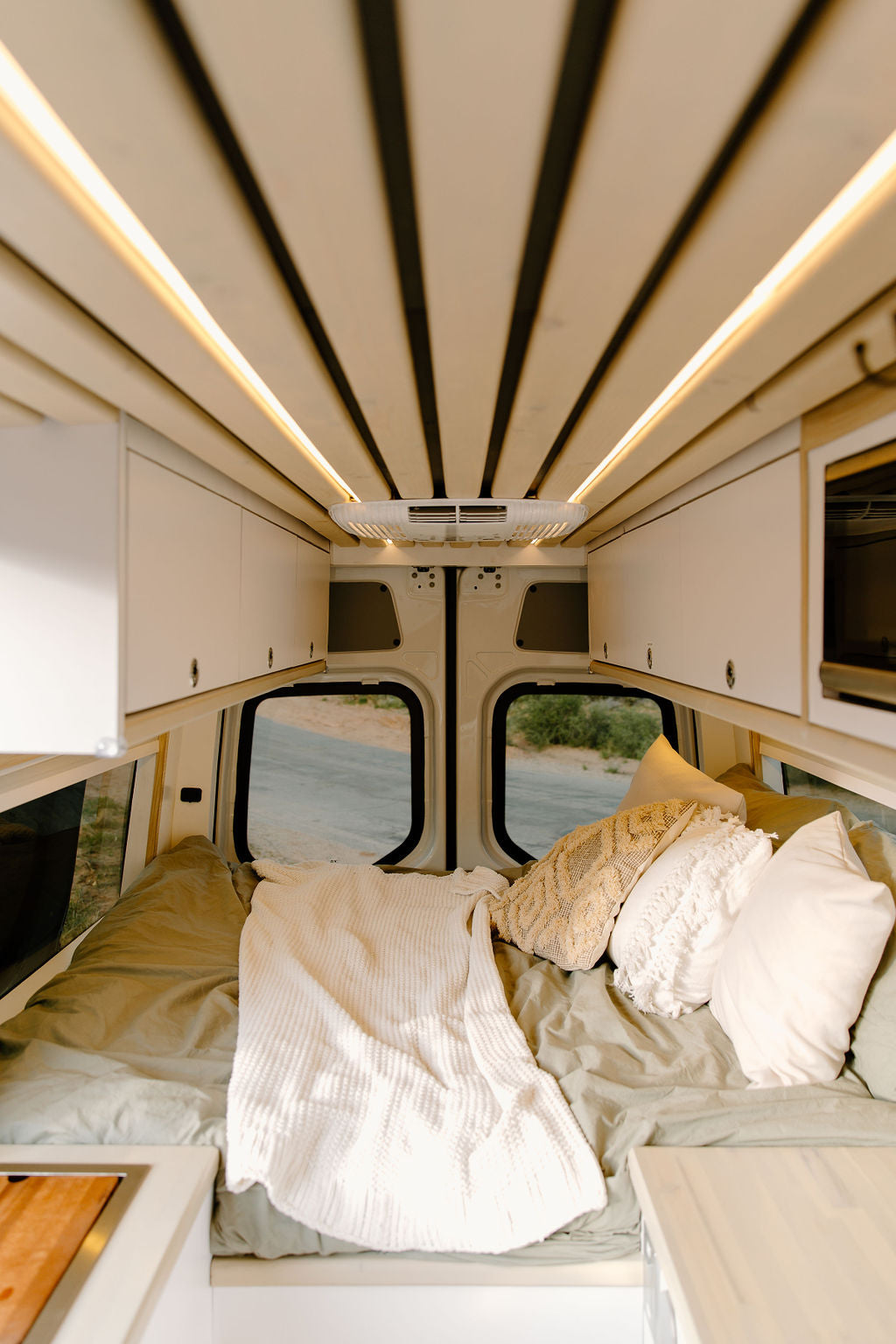 Inside a modern RV rental van: a cozy bed with pillows, sleek overhead cabinets, and ambient ceiling lights illuminating the space.
