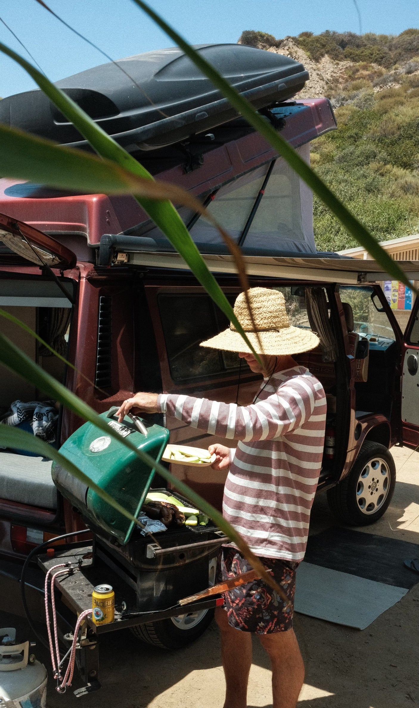 Man grilling beside a red pop-top camper van. An RV rental setup for outdoor cooking adventures.