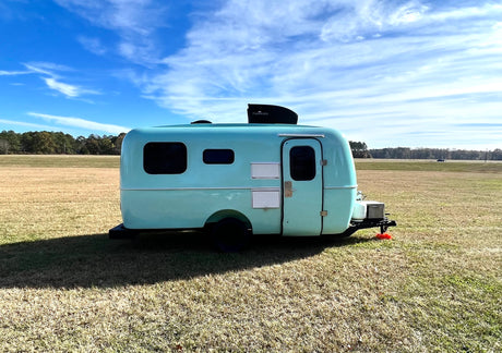Light blue vintage-style travel trailer (rv rental) with a Furrion unit, parked in a sunny field under blue sky.