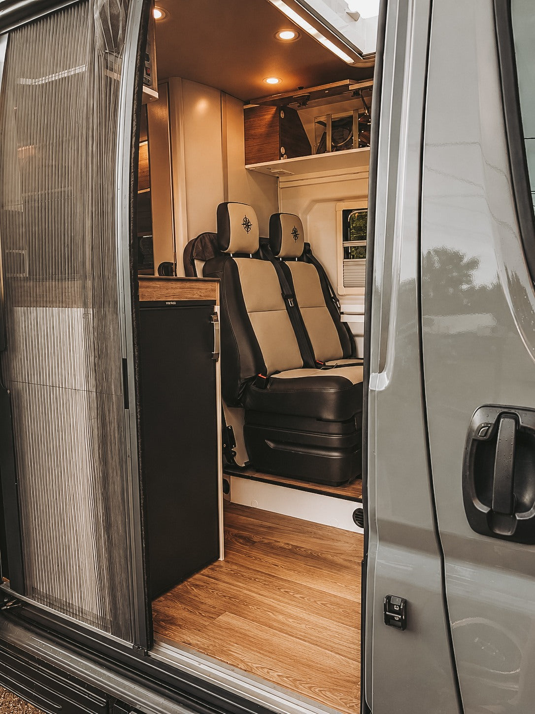 RV rental interior view: open sliding door reveals a cozy camper van with two-tone seating, wood floor, and fridge.