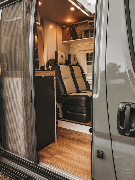 RV rental interior view: open sliding door reveals a cozy camper van with two-tone seating, wood floor, and fridge.