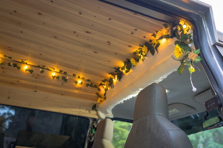 Cozy RV rental interior: wood-paneled ceiling with warm string lights and decorative foliage. Front seats visible, creating a charming camper feel.