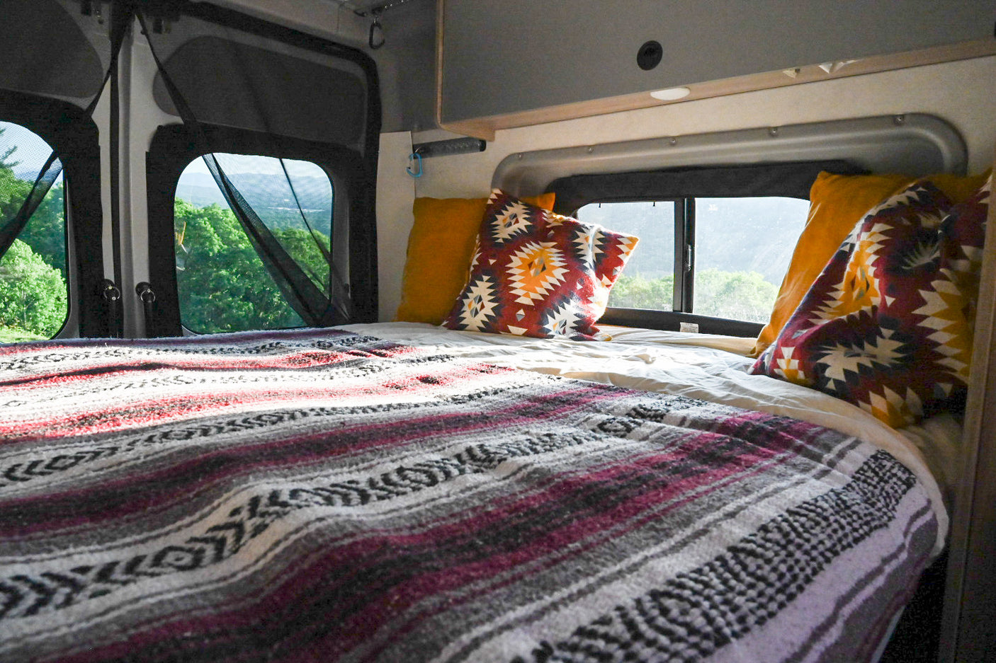 Cozy RV rental interior with bed, tribal blanket, and scenic forest views through netted windows.