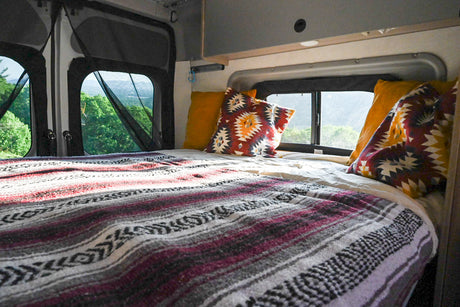 Cozy RV rental interior with bed, tribal blanket, and scenic forest views through netted windows.