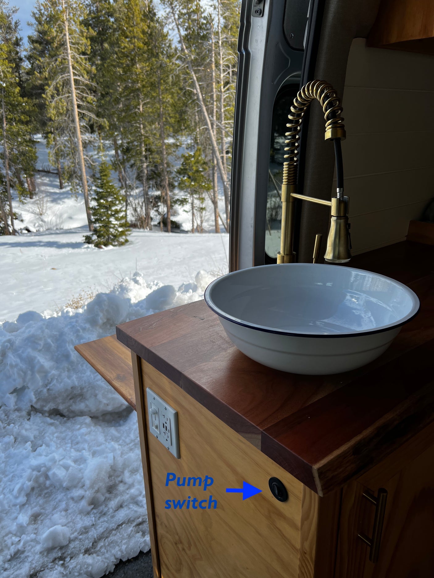 Interior of an RV rental: a modern kitchen sink with a golden faucet and water pump switch, overlooking a snowy forest.
