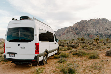 A white Mercedes Sprinter camper van RV rental parked in a vast desert landscape with rugged mountains under a cloudy sky.