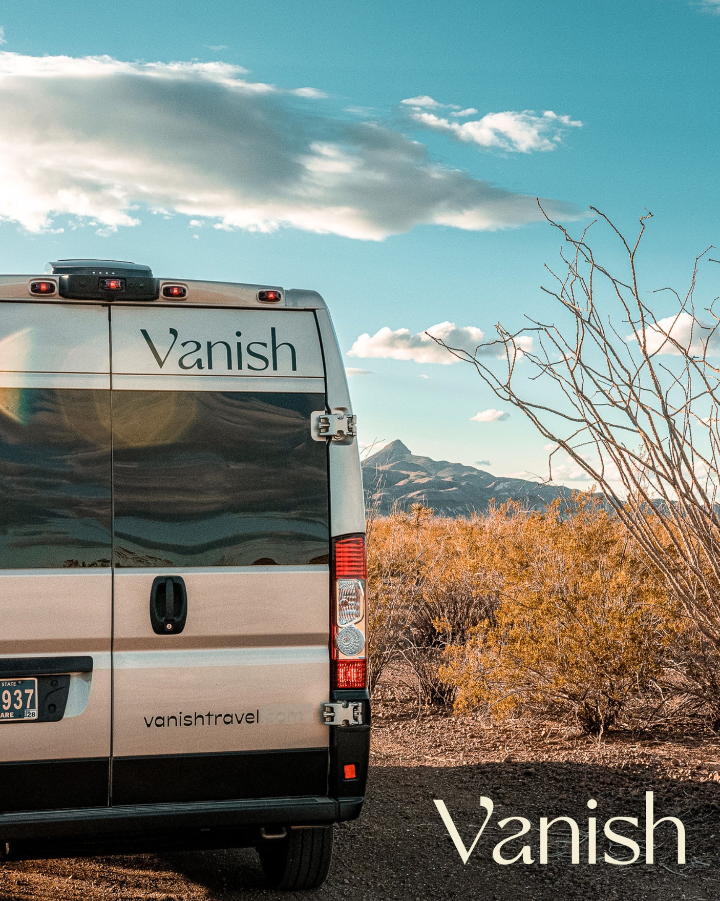 Rear view of a silver Vanish RV rental campervan parked in a desert landscape with mountains and blue sky.
