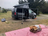 Enjoying an RV rental adventure! Grey pop-top camper van set up at a scenic campsite with solar power & firewood.