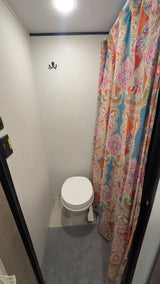 Inside an RV rental, a compact bathroom with a white toilet, vibrant paisley shower curtain, ceiling light, and wall hooks.