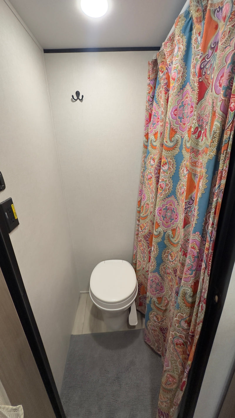 Inside an RV rental, a compact bathroom with a white toilet, vibrant paisley shower curtain, ceiling light, and wall hooks. — photo 7
