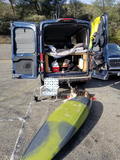 Blue rv rental van prepped for adventure: bed, gear, kayak, and a smiling dog. Ready for a great outdoor escape.