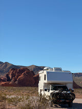 An RV rental with bikes on a rack, parked in a desert landscape with rugged red rock formations and mountains under a clear blue sky.