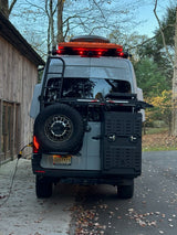 Rear view of an overland RV rental – a grey Sprinter van with red roof lights, ladder, spare tire, and gear storage boxes.