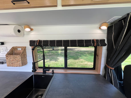 Cozy RV rental kitchen with a black sink, dark counter, window overlooking a green landscape, and striped valance.