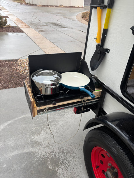 An RV rental's outdoor kitchen with a camp stove, pot, and frying pan ready for cooking. Axe & shovel mounted.