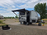 An RV rental with an extended awning is parked on a concrete pad at a campsite
