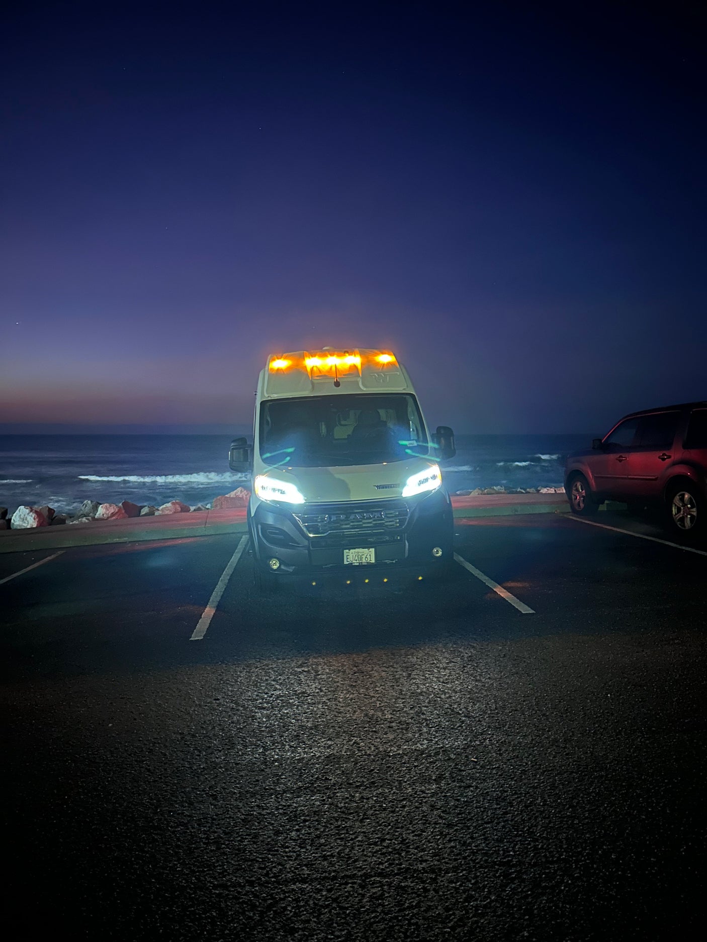 A white rv rental with bright headlights and amber roof lights is parked facing the camera, ocean and faint stars visible in the twilight.