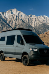 This grey off-road camper van RV rental is parked on dirt, with towering, rugged mountains and a clear sky for your adventure.
