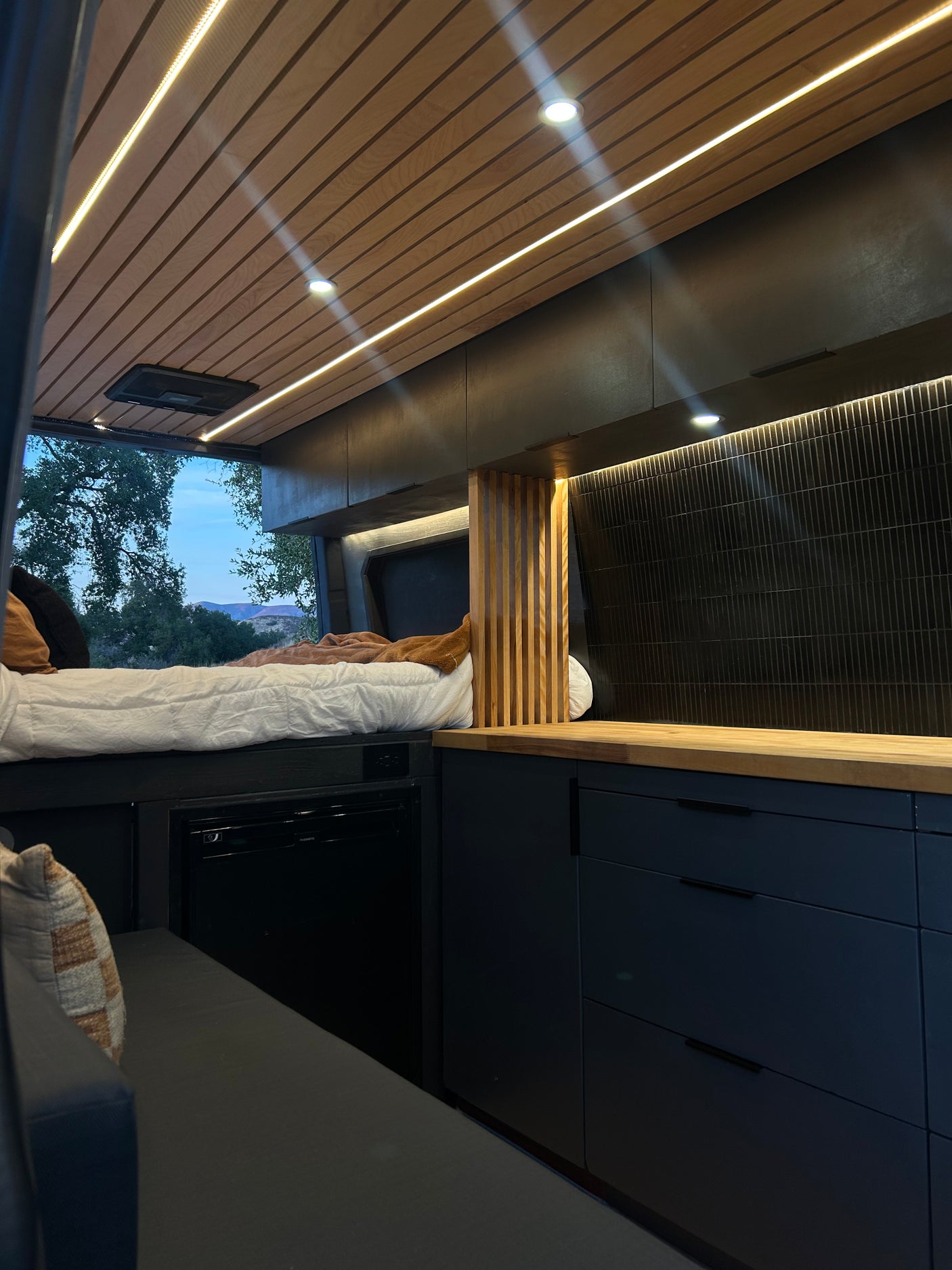 Modern RV rental interior: wooden ceiling with LED lights, cozy bed, compact fridge, dark cabinets, and nature view.