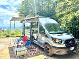 A silver Ford Transit RV rental with an awning and full outdoor camping setup, enjoying a beautiful mountain view.