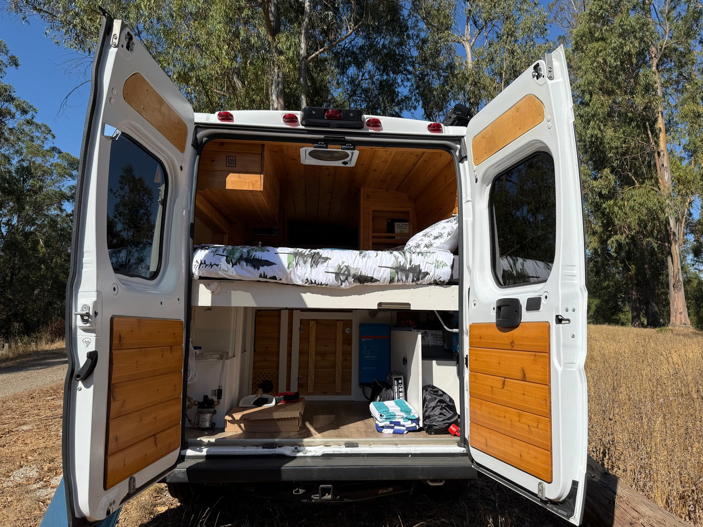 This RV rental showcases its custom-built interior, with open rear doors revealing a cozy bed, wood paneling, and power system. — photo 14