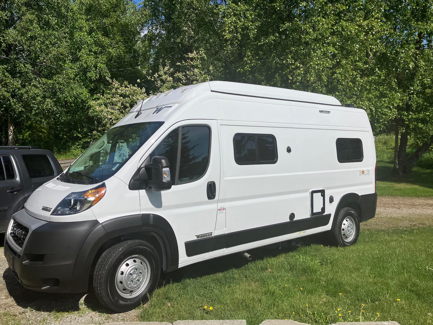 A white Winnebago Solis camper van, likely an RV rental, is parked on green grass with trees in the background.