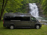 Grey rv rental camper van parked on grass, beside a river & grand waterfall. Lush green trees surround the stunning nature scene.