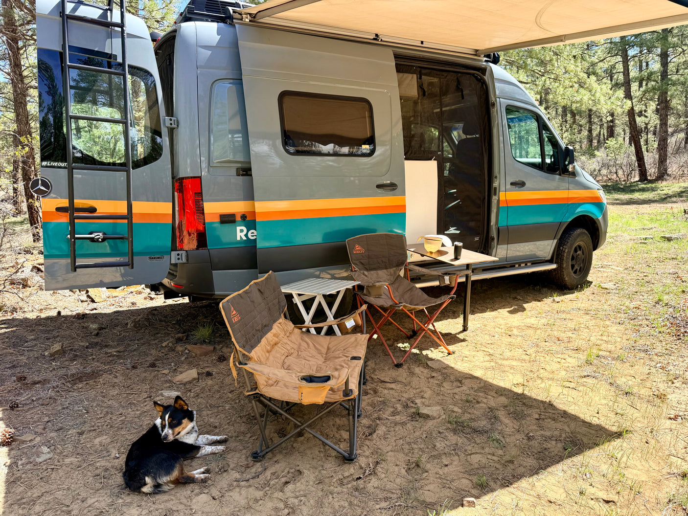 An rv rental camper van with an extended awning, outdoor chairs, and a dog relaxing at a scenic campsite.