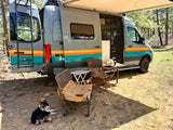 An rv rental camper van with an extended awning, outdoor chairs, and a dog relaxing at a scenic campsite.