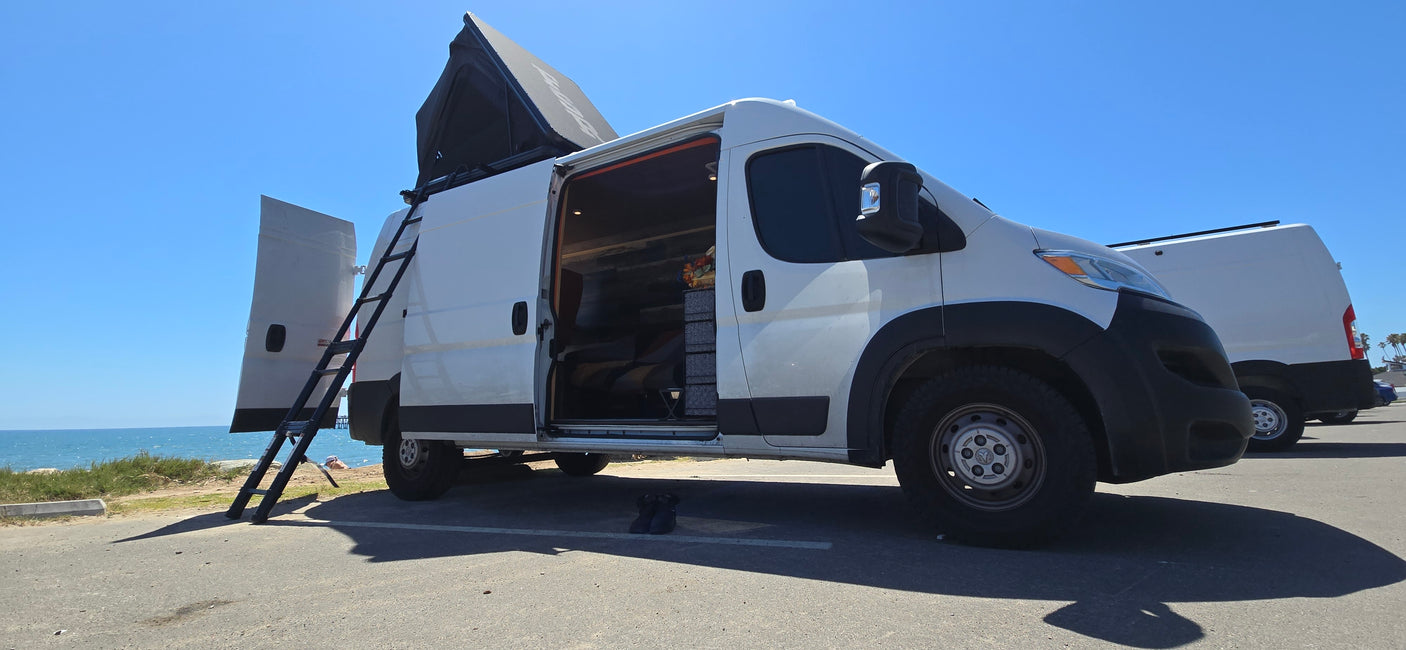 Coastal rv rental adventure: White camper van with pop-top open, ladder extended by the ocean on a sunny day.