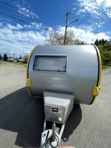 Front view of a sleek silver teardrop RV rental, featuring yellow trim and a storage box, parked on an asphalt road.