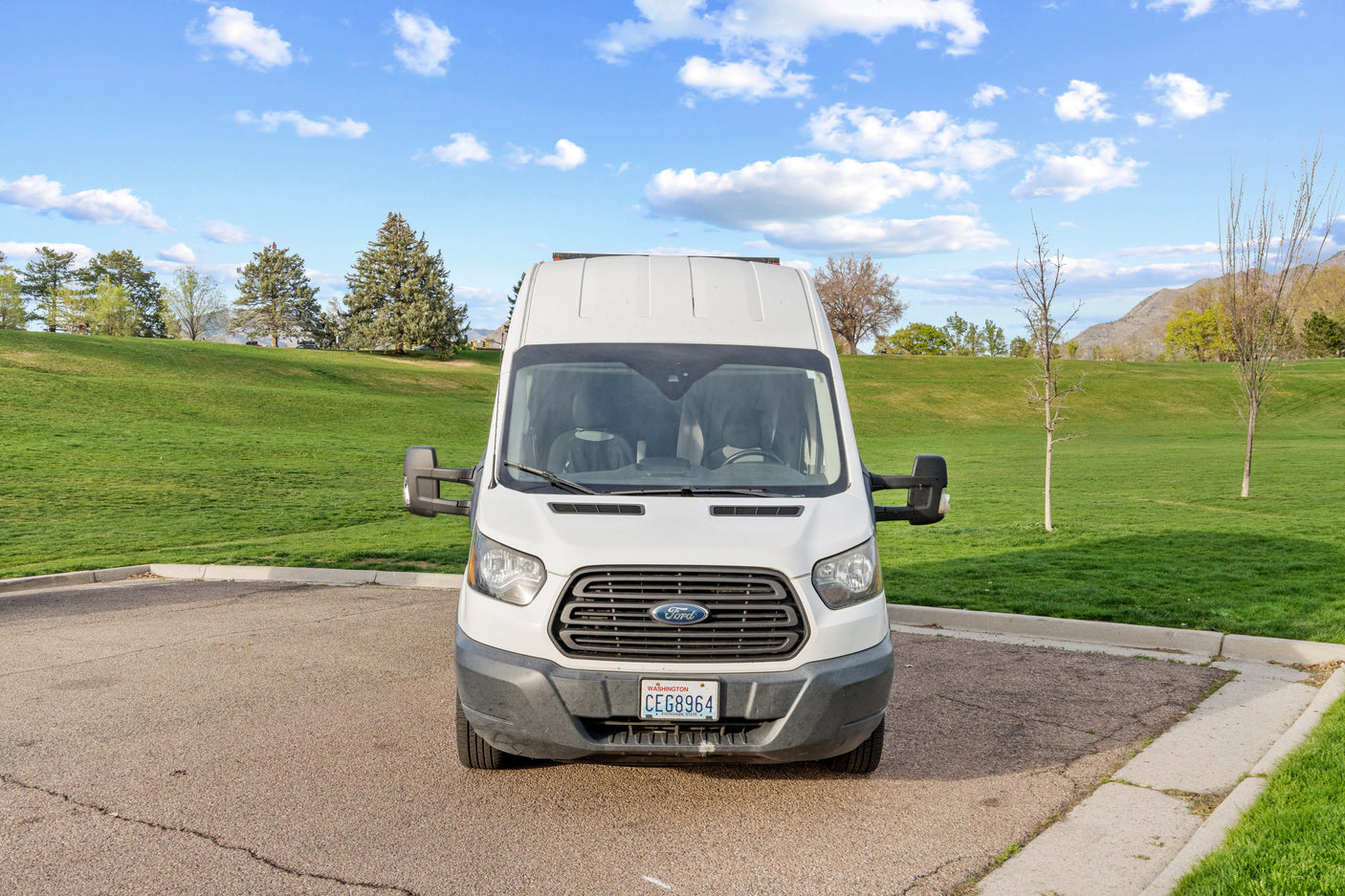 White Ford Transit rv rental van parked in a lot, backed by a green grassy hill, trees, and distant mountains.