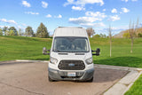 White Ford Transit rv rental van parked in a lot, backed by a green grassy hill, trees, and distant mountains.