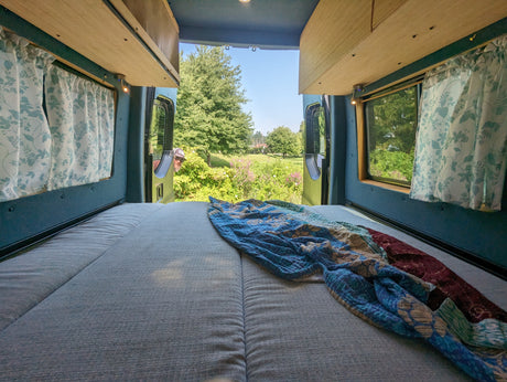 Relaxing RV rental interior looking out to nature. Bed, floral curtains, wooden shelves, and a person peering from outside.