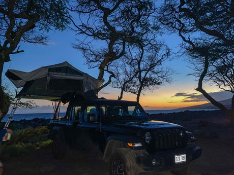 Experience nature with an RV rental: a Jeep Gladiator with a rooftop tent set up by the ocean at sunset.