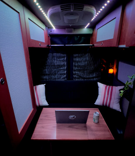This RV rental's cozy interior features a lit mobile workspace with laptop, seating, and blackout curtains.